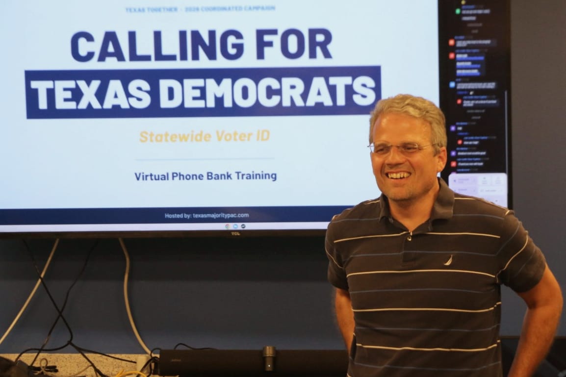 Smiling man with glasses stands in front of a large screen that reads'CALLING FOR TEXAS DEMOCRATS' during a training.