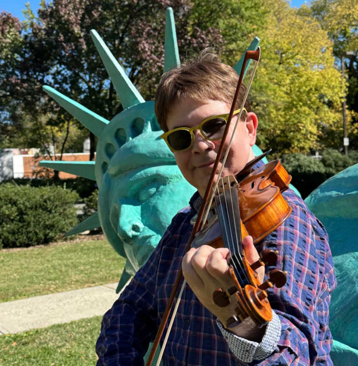 Leo Sushansky serenades the Reclining Statue of Liberty at Museum of Contemporary Art Arlington (photo by Steven Fleshman) | ARLnow Person wearing yellow sunglasses and a plaid shirt plays a violin outdoors, with a turquoise Statue of Liberty sculpture in the background.