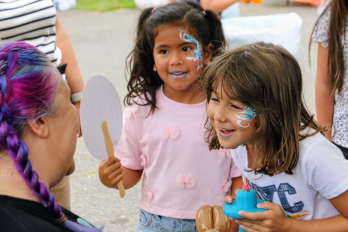 Two young girls with face paint gaze at a round handheld mirror held by a person with purple braided hair outside. (Informative)