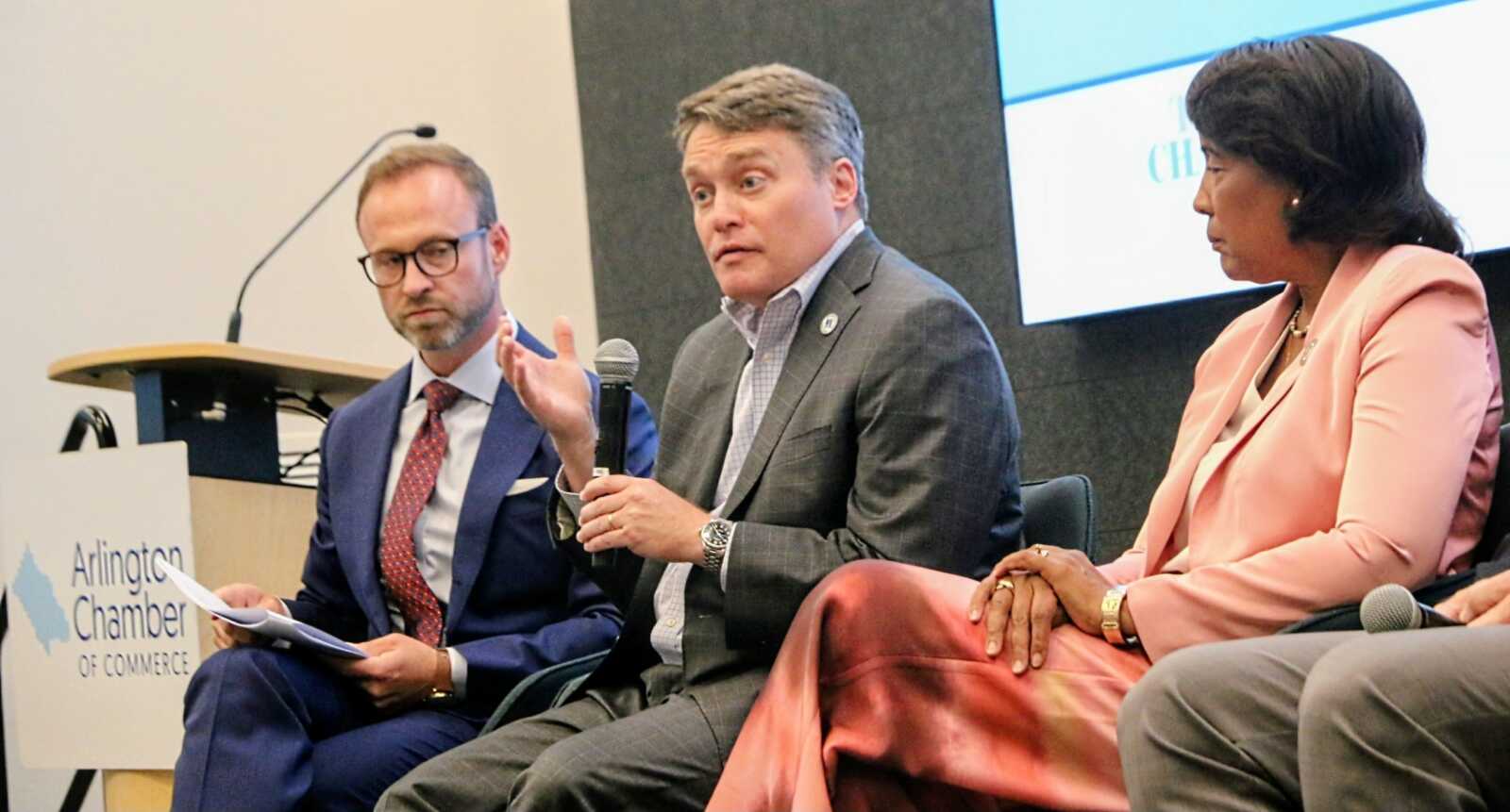 Panel discussion at Arlington Chamber of Commerce with three professionals, one speaking into a handheld mic while two others listen nearby, on a stage with a lectern and sign visible on the left.