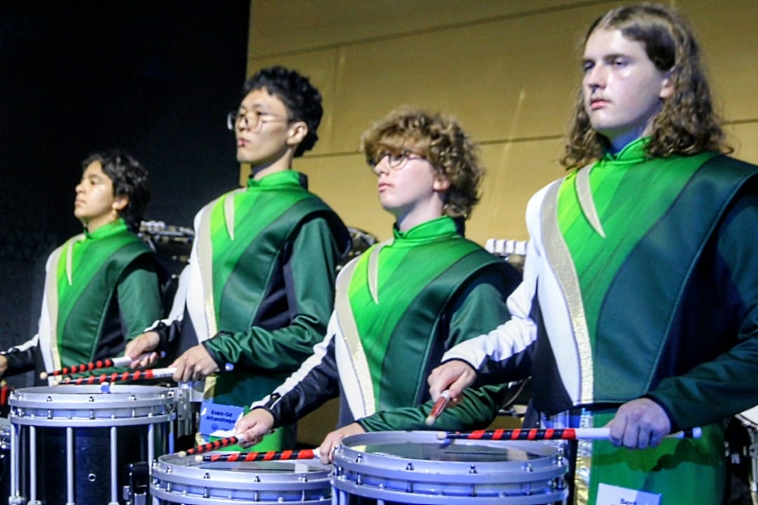 Four young percussionists in matching green uniforms playing marching snare drums on a stage.