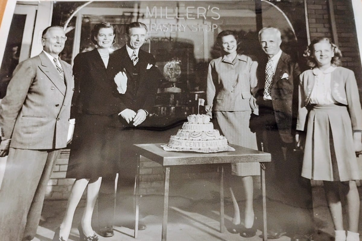 Six adults in vintage attire pose around a table with a multi-tier centennial cake outside Miller's Pastry Shop.