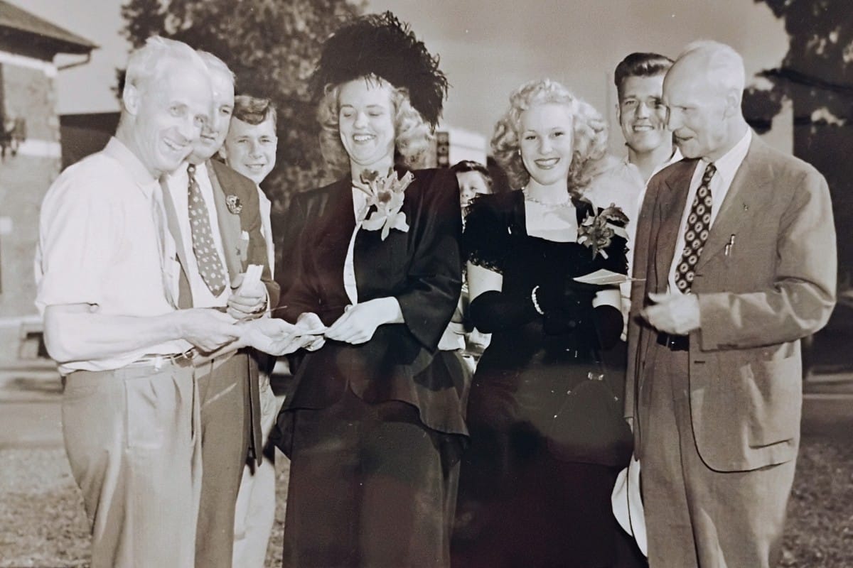 Group of elegantly dressed people outdoors, two women in formal gowns exchange a card or envelope with men smiling nearby.