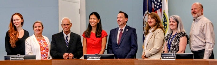 Eight adults stand behind a long wood-paneled table in a formal meeting room, smiling, with flags visible on the right.