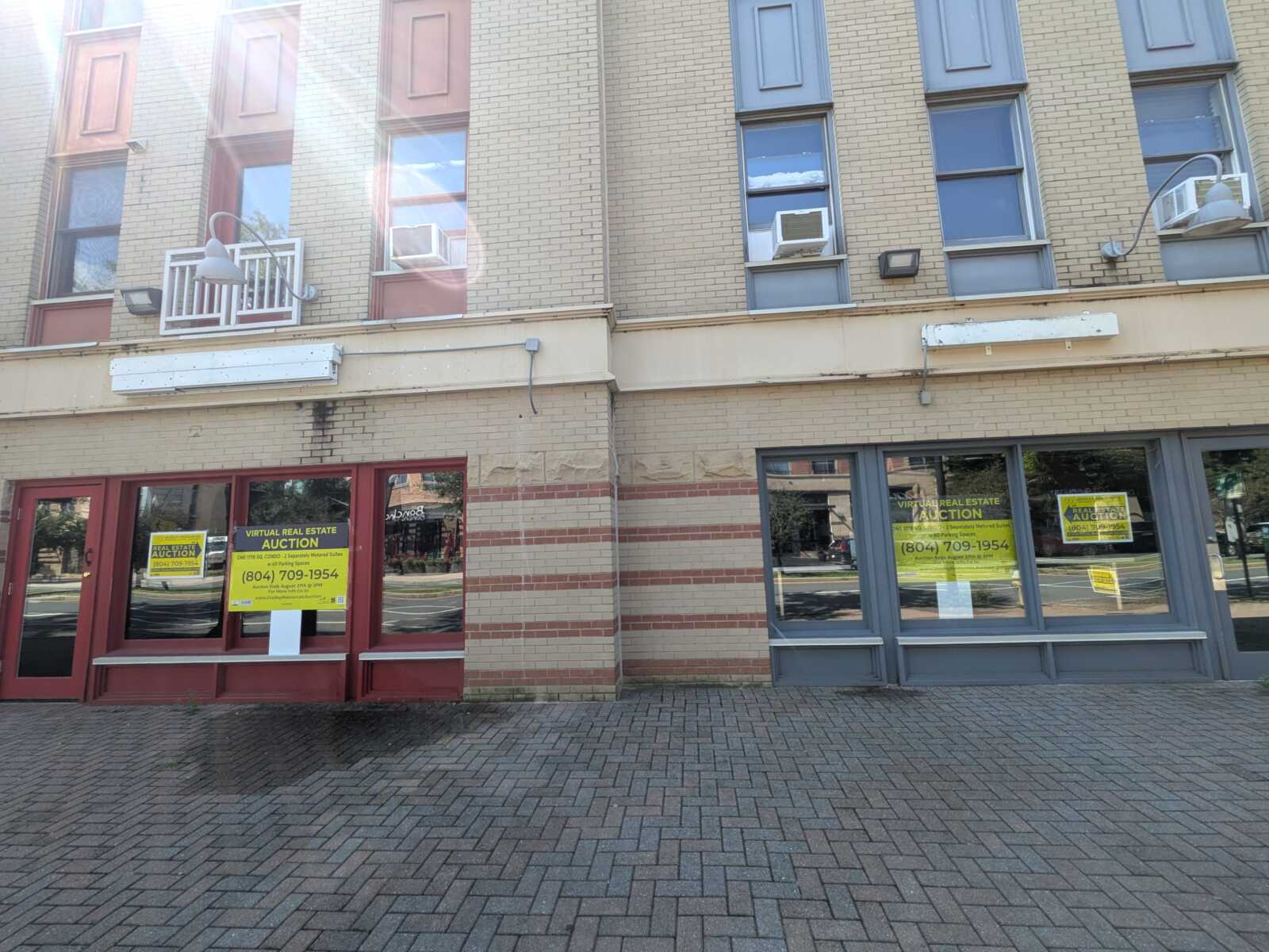 Street view of a brick storefront building with large windows and yellow auction signs in the windows, advertising a virtual real estate auction with a phone number.