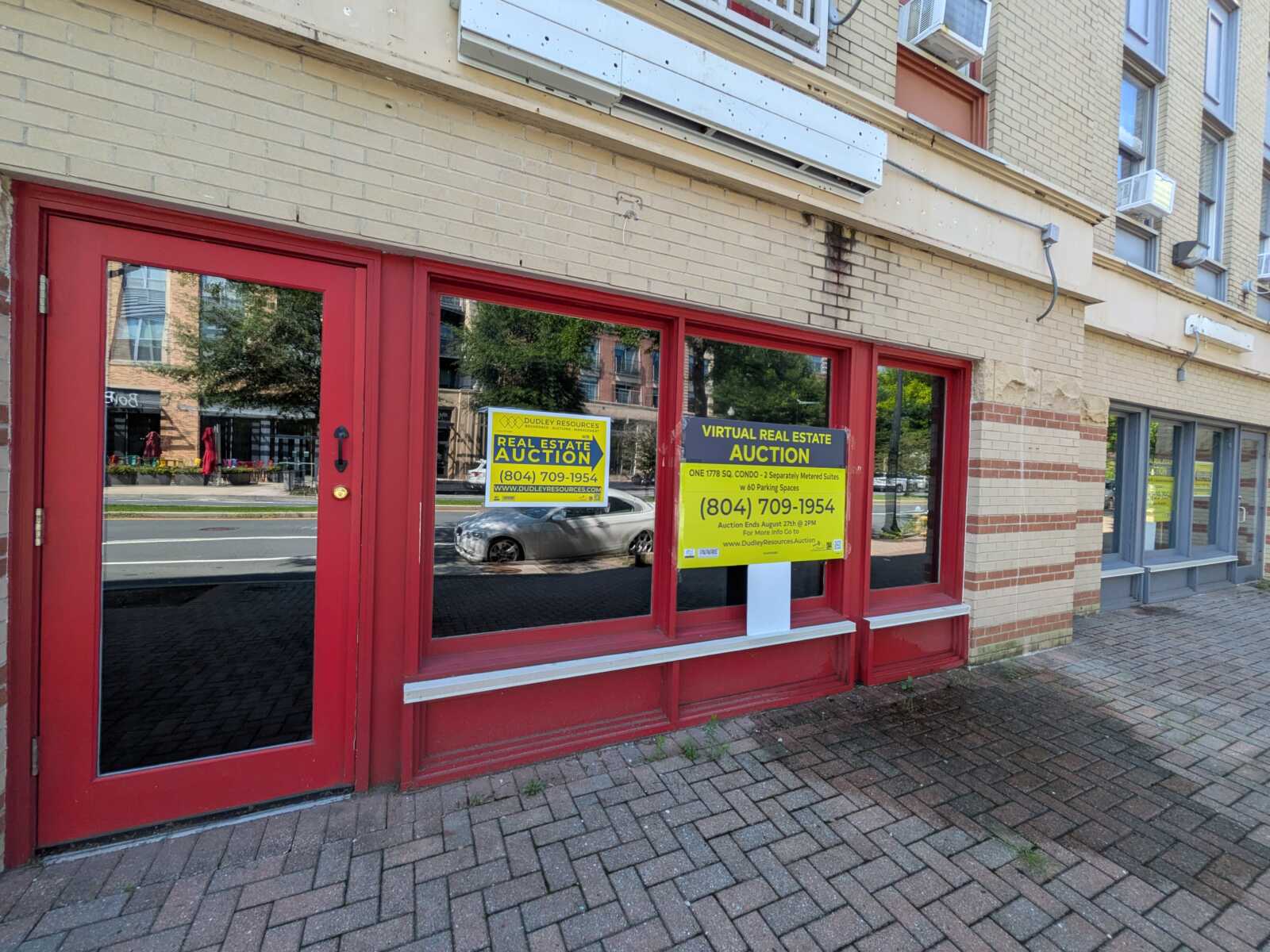 Red-framed storefront with large glass windows and two bright yellow auction signs reading 'Real Estate Auction' on the windows.