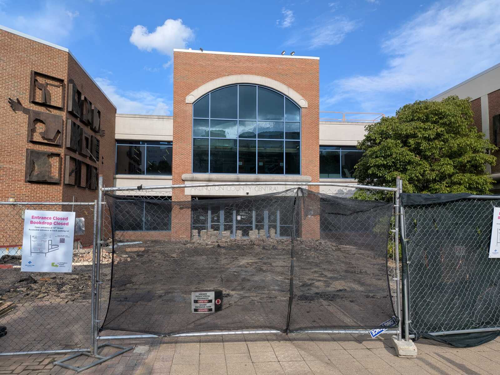 Construction fence blocks the brick building entrance undergoing renovation; signs say Entrance Closed and Bookdrop Closed.