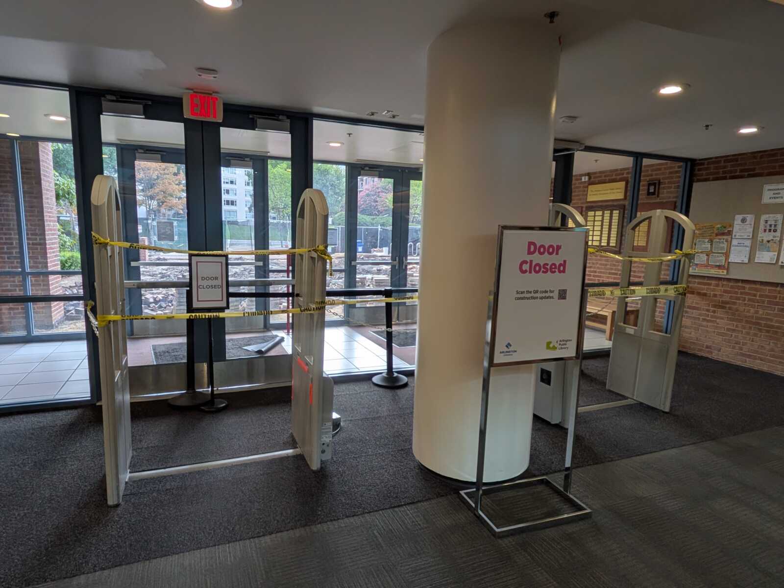 Entrance area with yellow caution tape across turnstiles and a 'Door Closed' sign; glass doors and an exit sign visible in a library or lobby.
