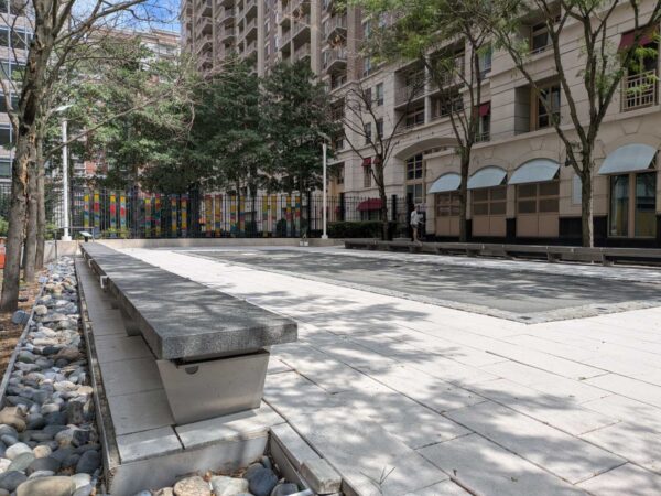 Urban plaza with long concrete benches, pebble border, and a colorful fence near a beige apartment building.