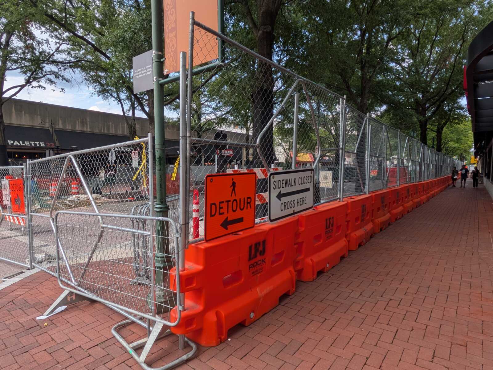 Construction barriers line a city sidewalk with orange detour sign and 'Sidewalk Closed' message; pedestrians in the distance.