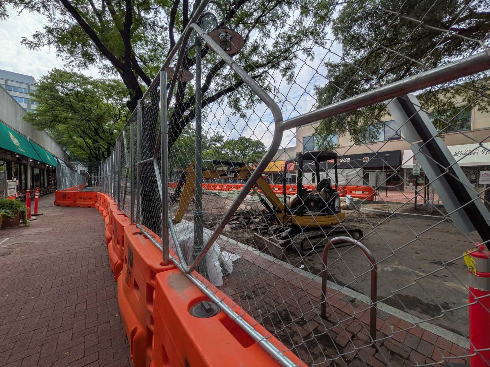 Construction site on a city street fenced off with orange barriers and a small excavator behind it.