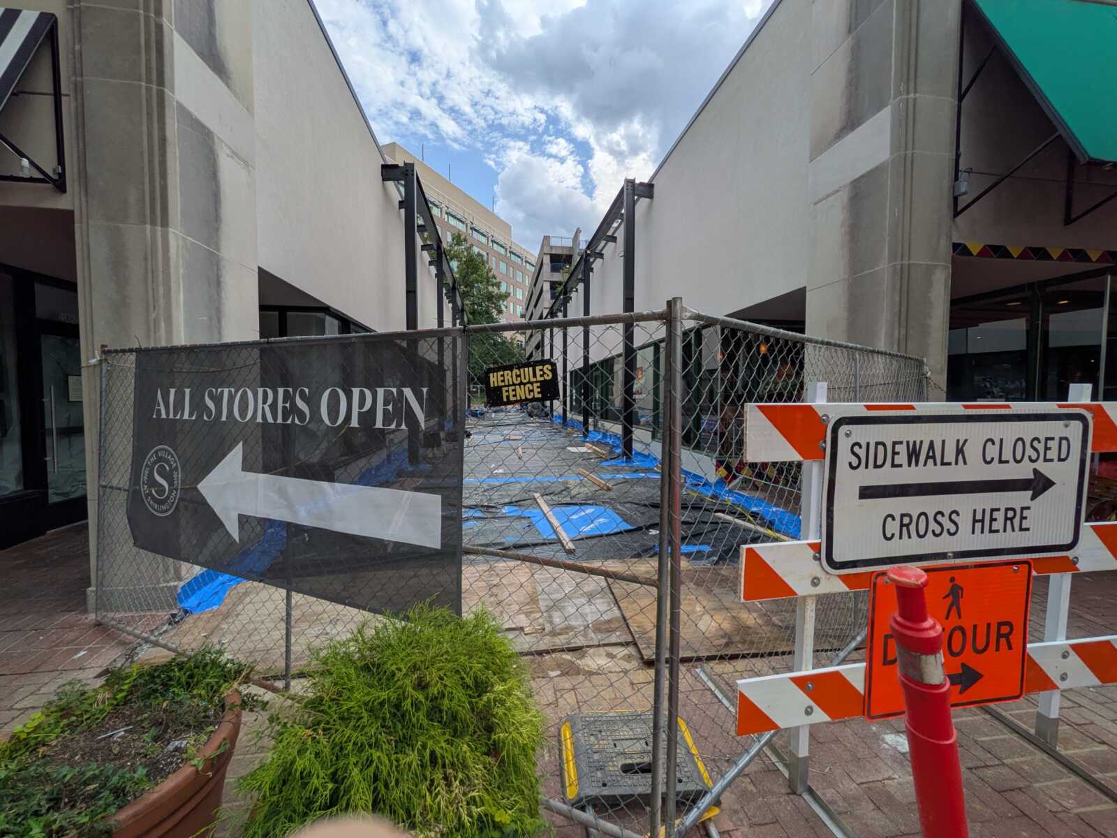 Construction fencing blocks the walkway between buildings; signs indicate 'All Stores Open' and 'Sidewalk Closed' nearby.