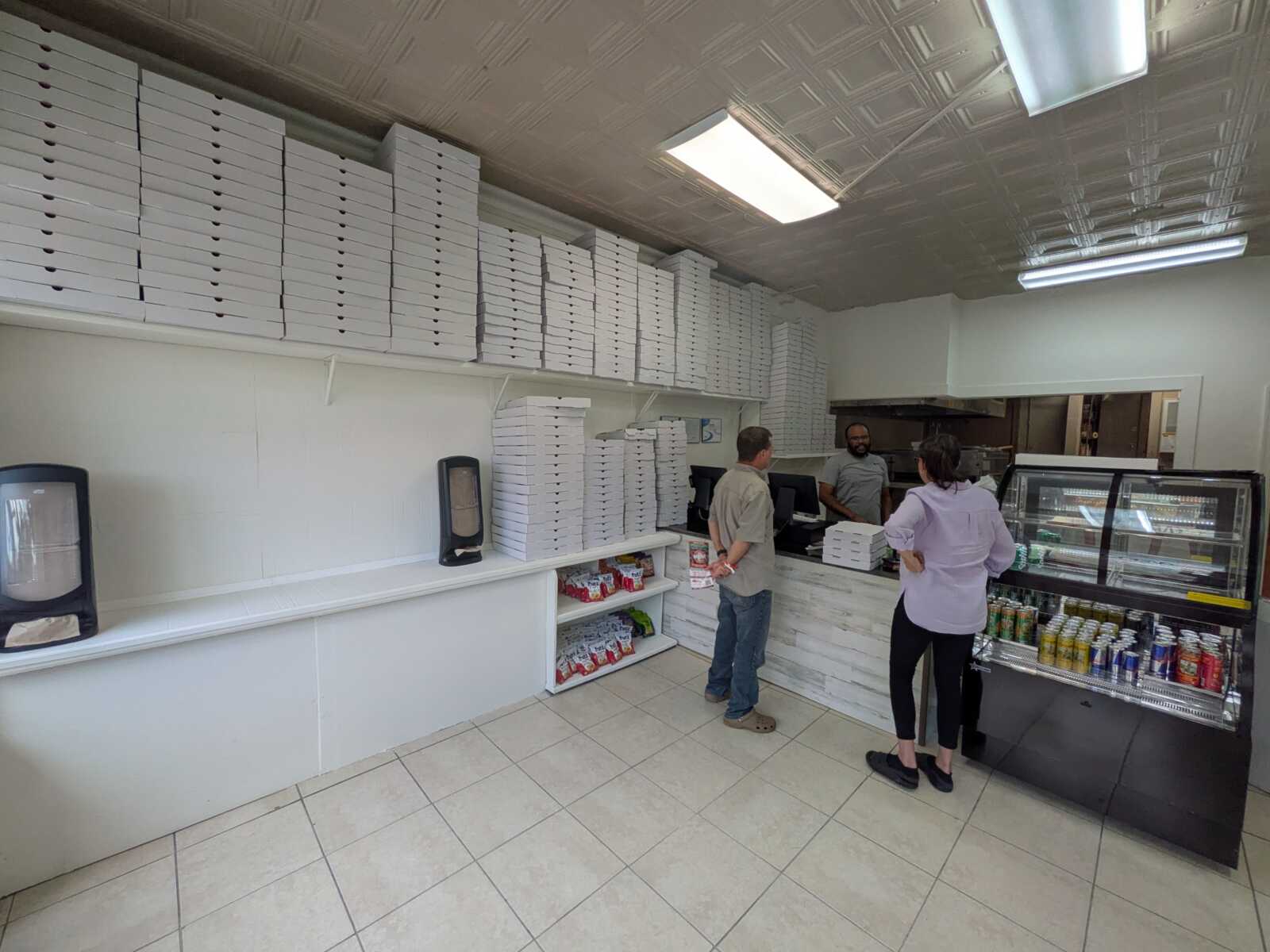 Pizza shop counter with tall stacks of white pizza boxes along the wall and staff behind the counter.