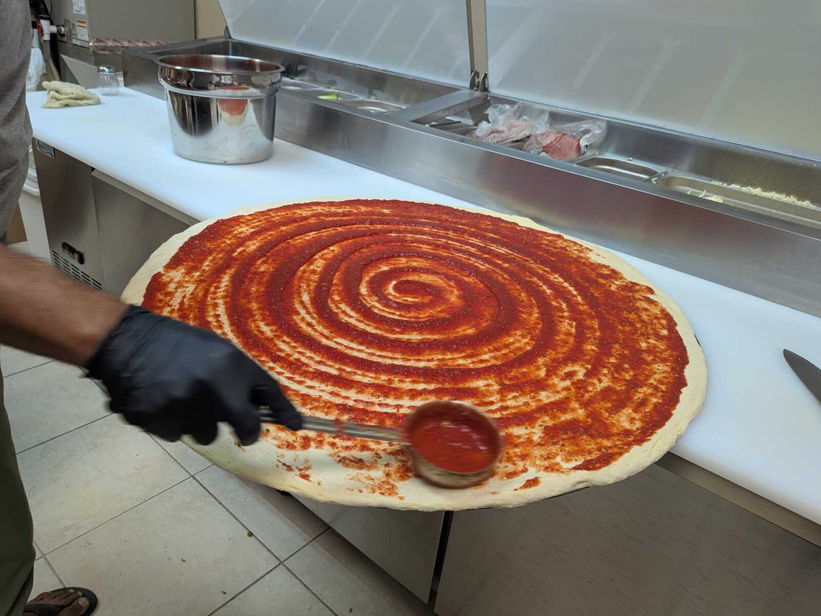 Large round pizza dough on a prep table with a red tomato sauce spread in a spiral pattern, a chef ladling sauce onto it in a professional kitchen.