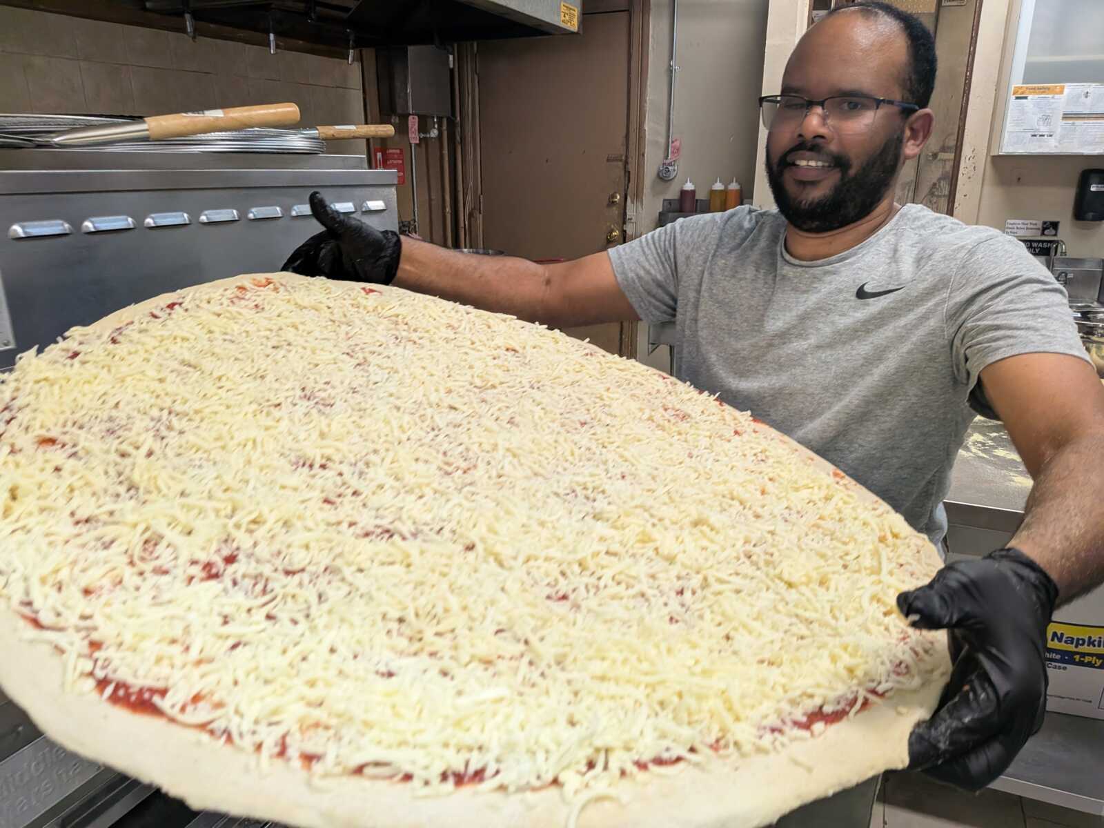 Smiling man wearing a gray t-shirt and black gloves holds a giant cheese pizza in a kitchen.