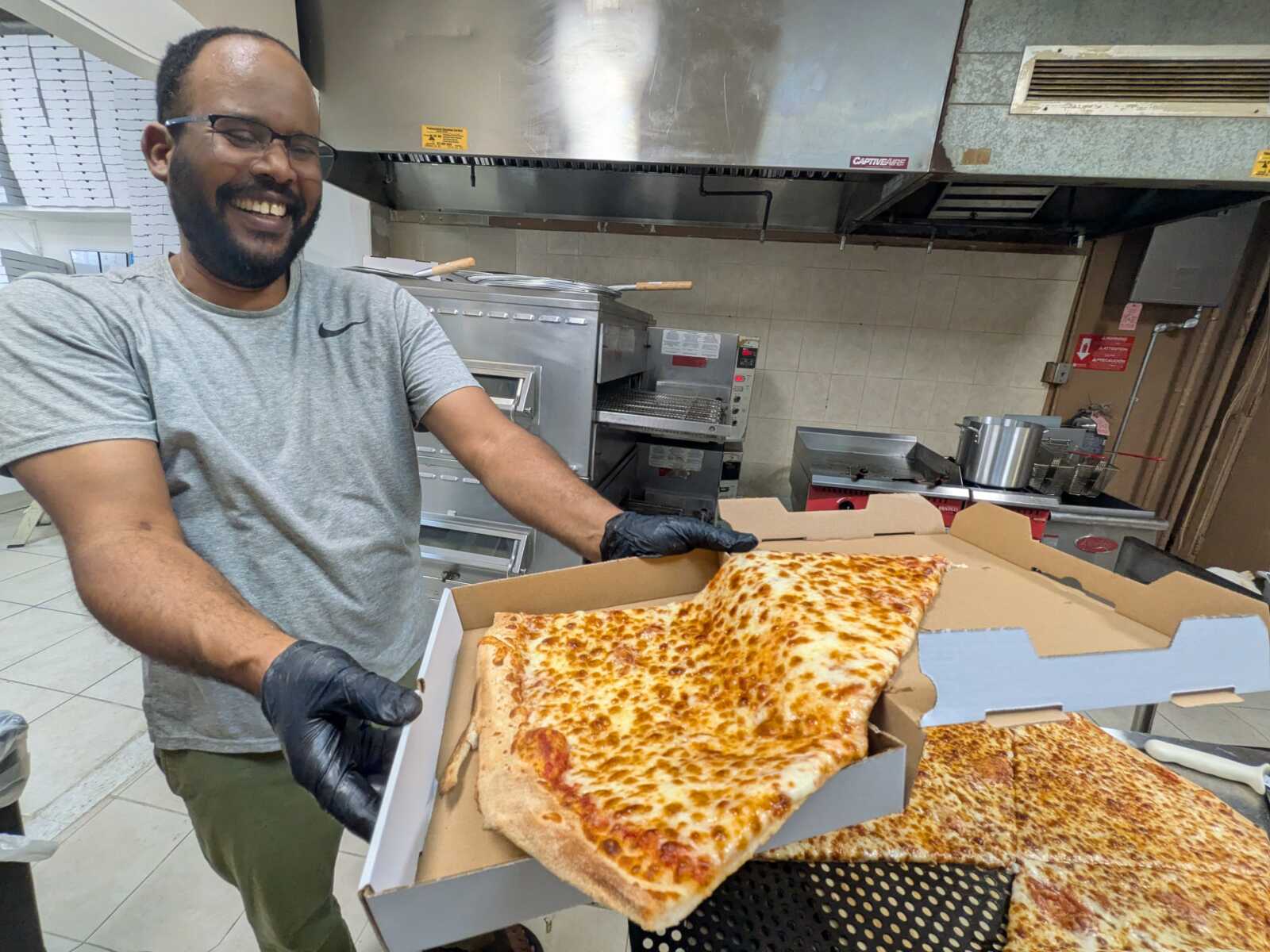 Smiling staff member in glasses and gloves presents a cheese pizza in a commercial kitchen, holding an open pizza box.
