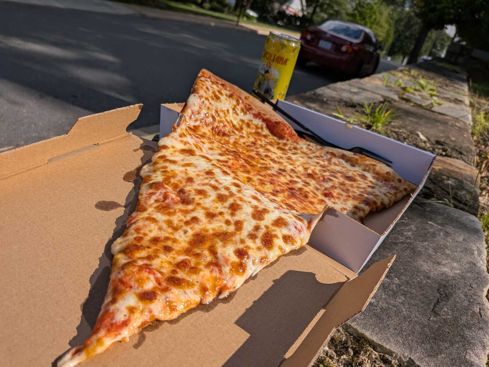 Large cheese pizza slices in an open pizza box placed on a concrete surface beside a street, with a yellow can in the background.