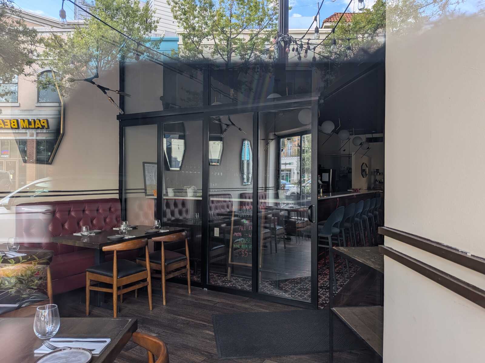 Restaurant interior with red tufted booths, wooden chairs, and tables set with wine glasses seen through glass storefront doors.