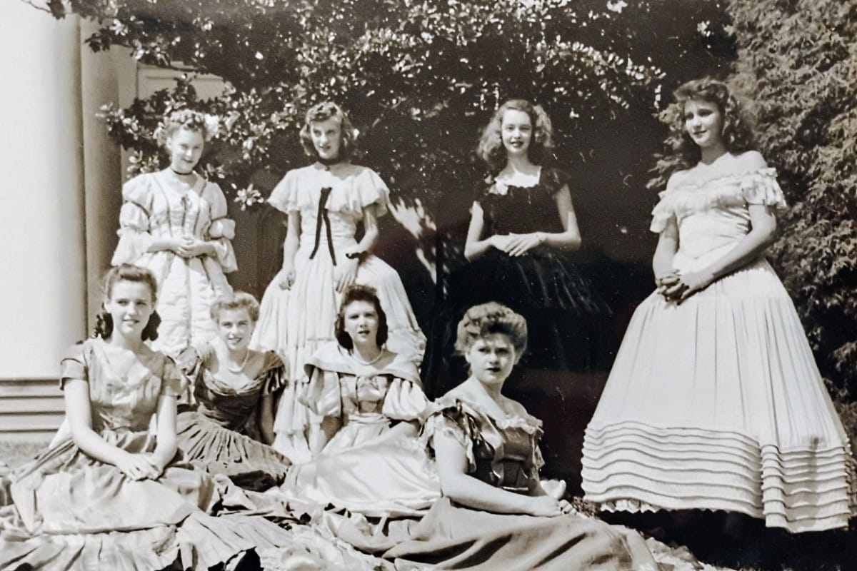 Group of seven young women in ornate 1800s-style dresses posing outdoors in a garden setting.