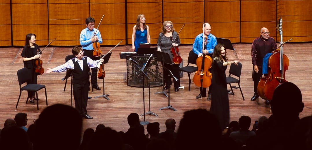 Performing Vivaldi’s The Four Seasons with with guest host-narrator Lynda Carter (photo by David Moss) | ARLnow A small orchestra on a wooden stage with violinists, a cellist, and a keyboardist; a young boy in front appears to conduct or lead while a woman in black raises her arms toward the audience, with audience silhouettes in the foreground.