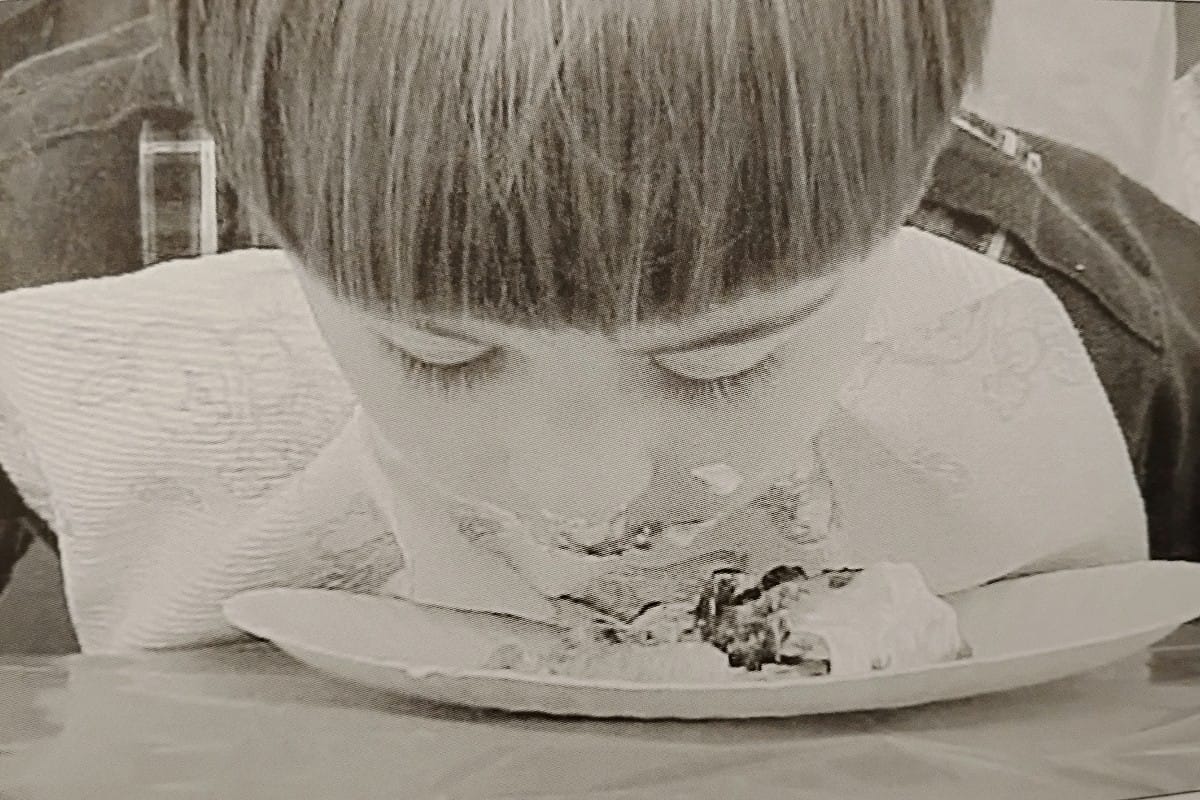 Young child leans over a plate, eyes closed, with frosting on their nose as they eat cake.