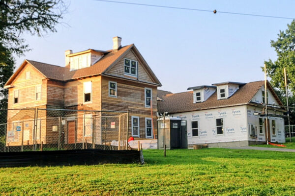 A two‑story house under renovation: one side with exposed wood siding and windows, the other wrapped in Tyvek with a portable toilet nearby outside a chain‑link fence.
