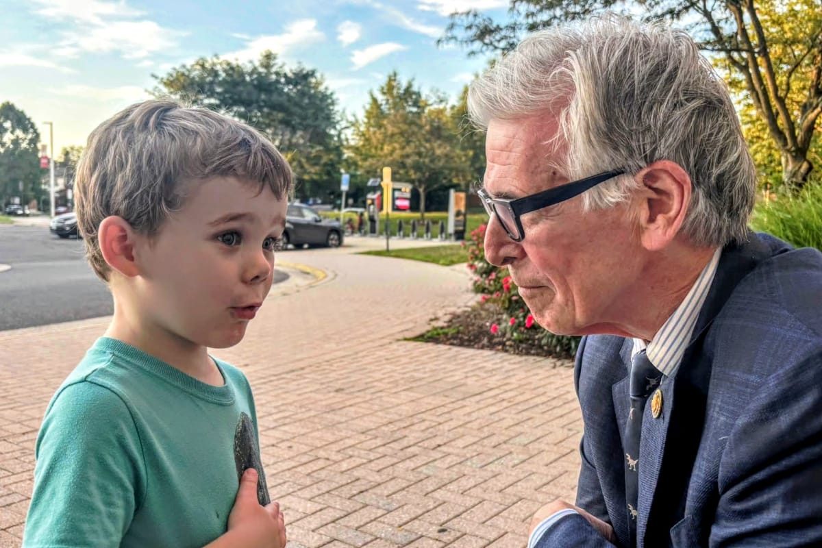 Boy in a teal shirt talks with an older man in a blazer and glasses on a brick sidewalk outdoors, near a street.