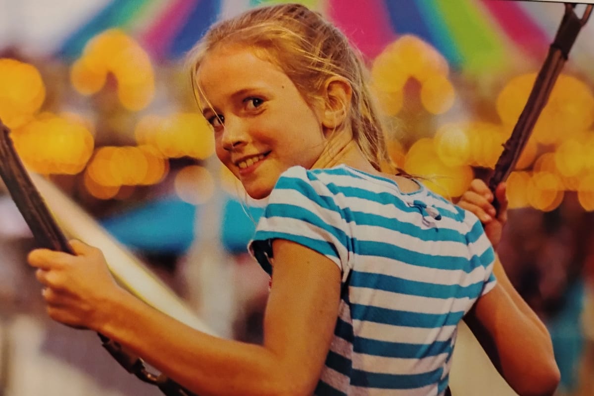 Smiling girl in a blue-and-white striped shirt holding ropes on a carnival swing ride, colorful lights in the background.