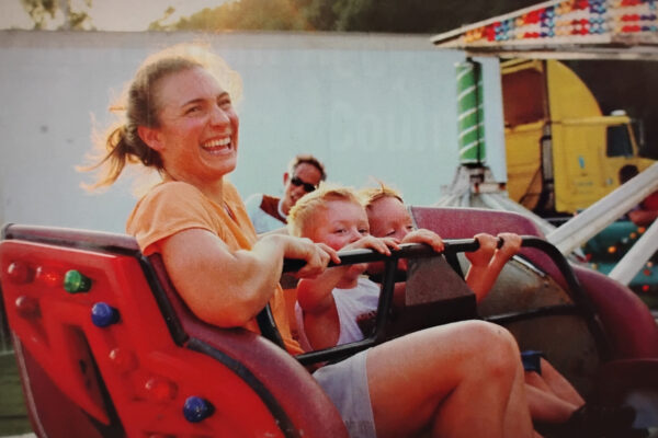 Woman laughing on a red amusement ride with two children beside her, sunny day at a fairground.