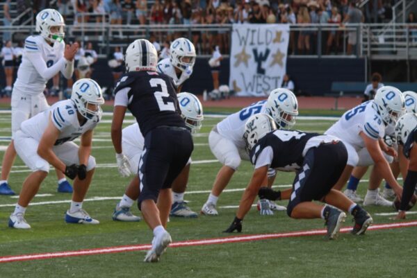 Football game at the line of scrimmage: white-uniformed offense facing a dark-uniform defender, players crouched and ready to snap, with fans and a 'WILD WEST' banner in the background.