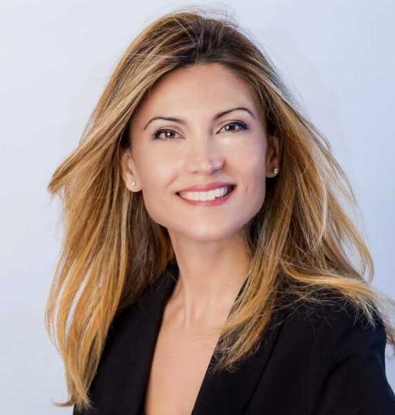 Professional woman with long blond hair in a black blazer, sitting at a table and smiling at the camera.