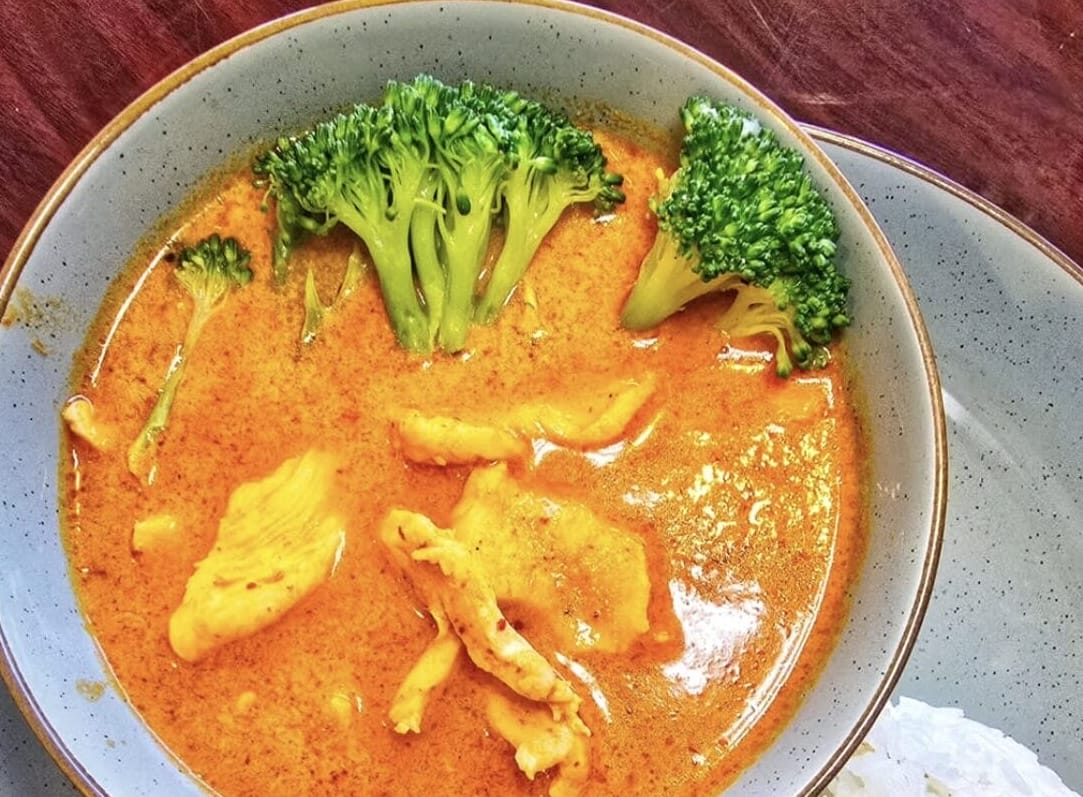 Bowl of orange curry soup with broccoli florets and chicken pieces, served with rice on the side.