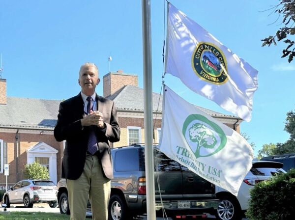 Older man in a blazer stands outdoors near flagpoles with the City of Falls Church, Virginia flag and an Arbor Day Foundation flag behind him on a sunny day in a parking lot.