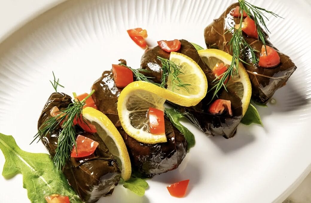 Plated stuffed eggplant rolls garnished with lemon slices, dill, and red peppers on a white dish.