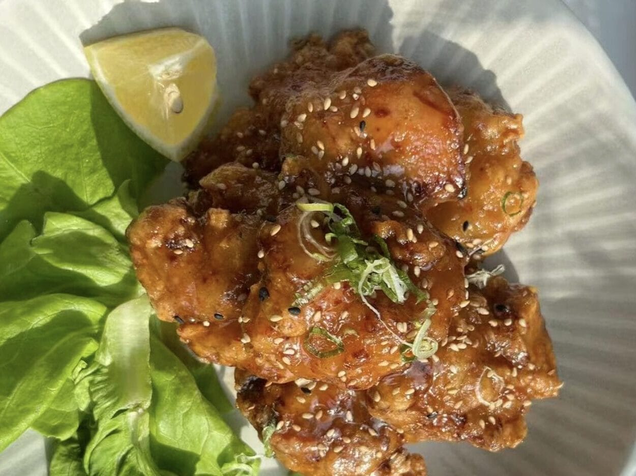 Crispy fried chicken pieces glazed with sauce, sprinkled with sesame seeds, on a plate with lemon and greens.