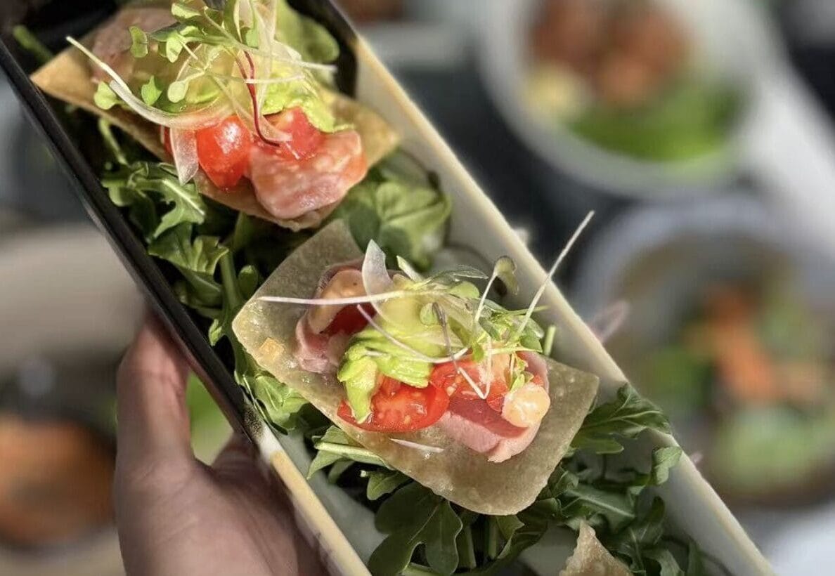 Two small crispy tortilla cups filled with greens, chopped tomatoes, and microgreens, held in a hand on a serving tray.