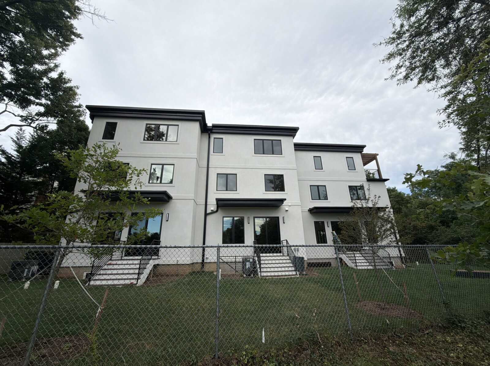Three-story white modern townhouses with black trim behind a chain-link fence on a cloudy day.