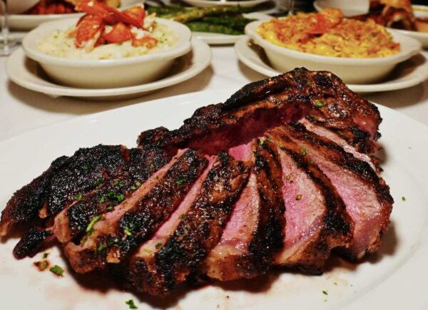 Sliced barbecued ribs on a white plate, dark crusted exterior and pink smoke ring visible, with side dishes in the background.