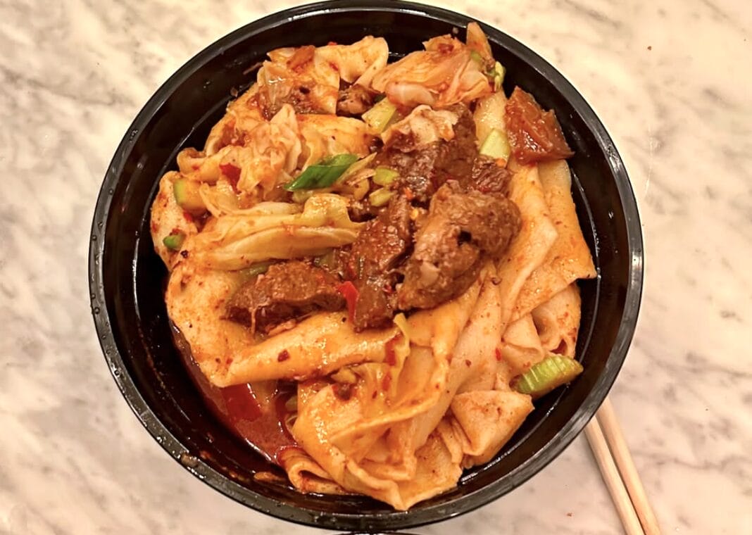 Black bowl of spicy Korean noodle soup with beef, kimchi, and green onions on a marble table; chopsticks rest beside the bowl.