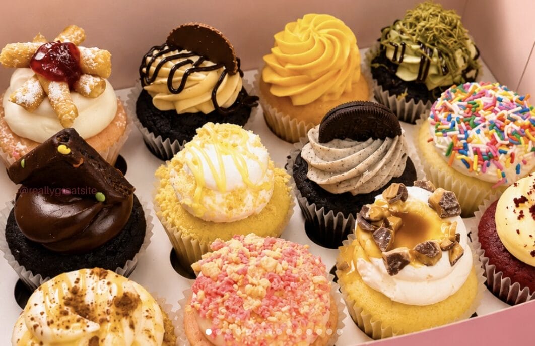 Assorted cupcakes in a box with colorful swirled frosting and toppings like sprinkles, cookies, and fruit pieces.