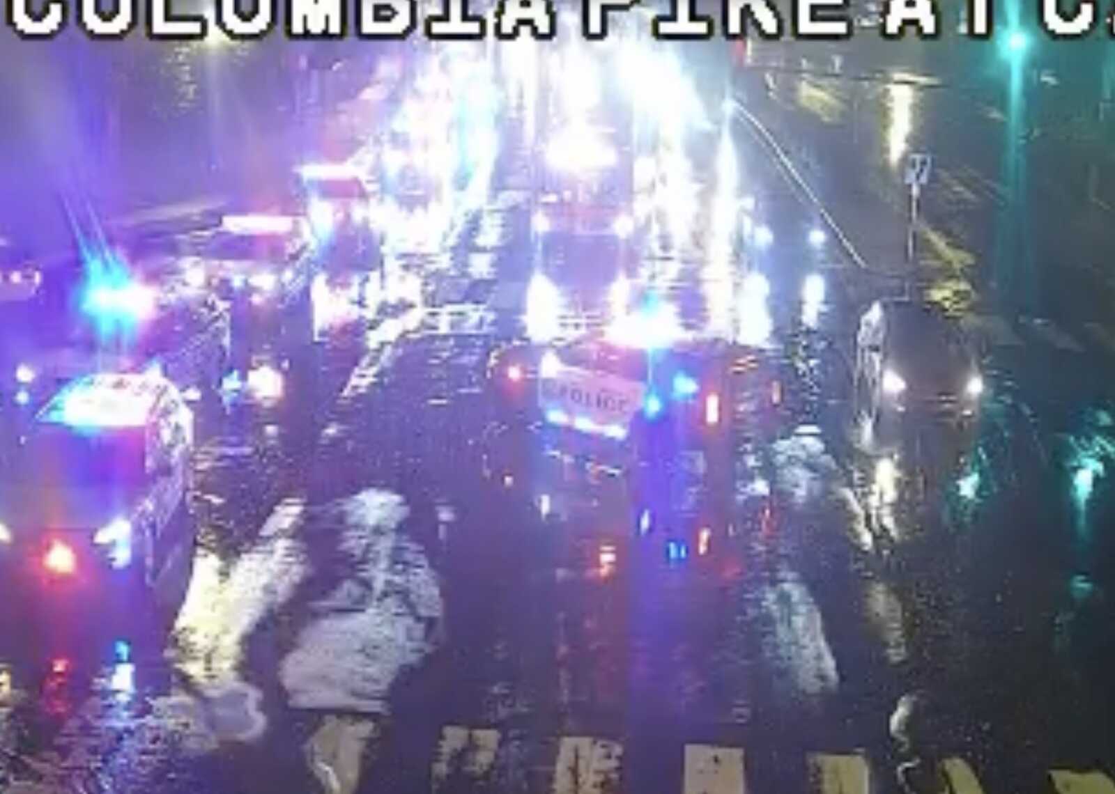 Nighttime city street with multiple police cars and flashing red/blue lights in the rain; active emergency response scene.