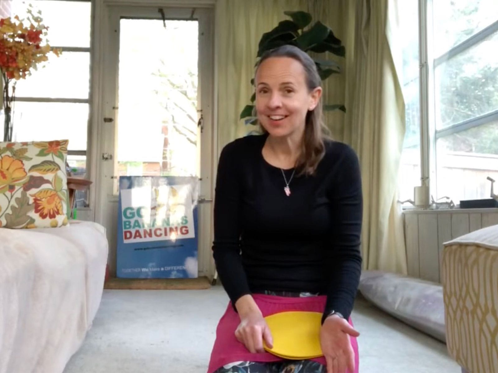 Woman sits on a light carpet in a bright living room, smiling at the camera while holding a pink and yellow circular item in her lap; colorful pillow and a poster are visible in the background.