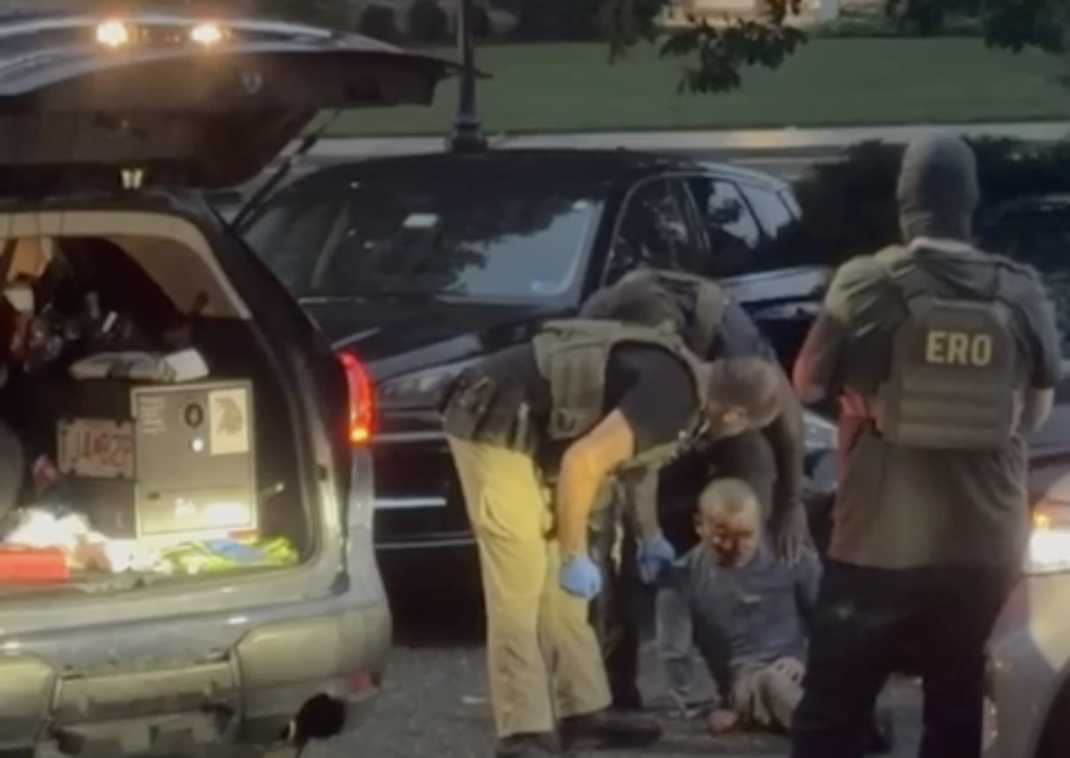 Tactical officers in vests arrest a man sitting on the ground beside a black SUV with the trunk open.