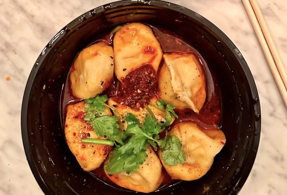 Black bowl of dumplings in red chili sauce topped with cilantro and sesame seeds, chopsticks beside the bowl