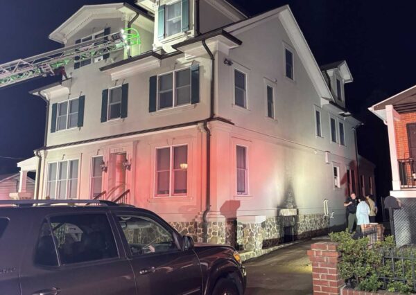 Night scene of a white two-story house at night with a fire truck ladder extending to the roof and pink emergency lighting on the facade; a few people stand near the entrance.