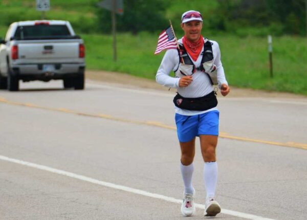 Runner wearing a white long-sleeve shirt, blue shorts, and a red bandana runs along a rural road with an American flag attached to his vest; a pickup truck behind him.