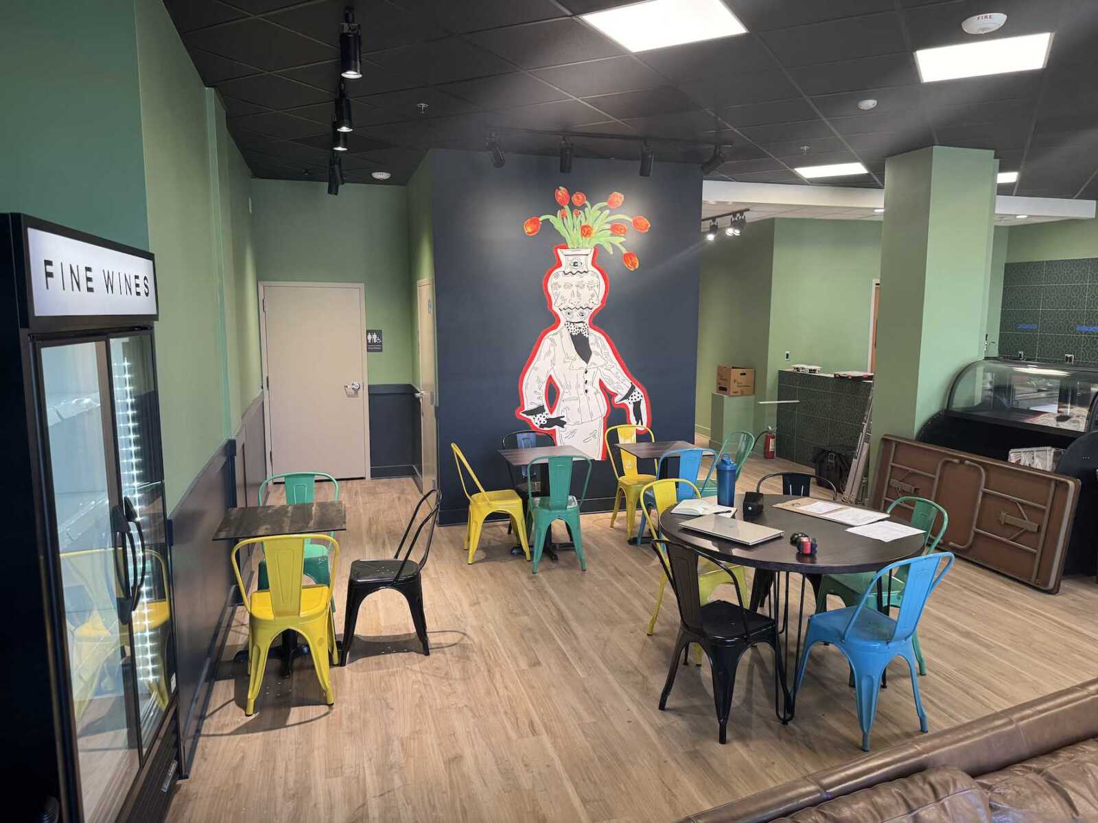 The interior of Good Trouble Roasters (courtesy of Mike Greiner) | ARLnow Indoor cafe area with colorful metal chairs around round tables, a large mural of a white-clad figure with a vase of red flowers on its head on a blue wall, and a wine fridge labeled 'FINE WINES' on the left.
