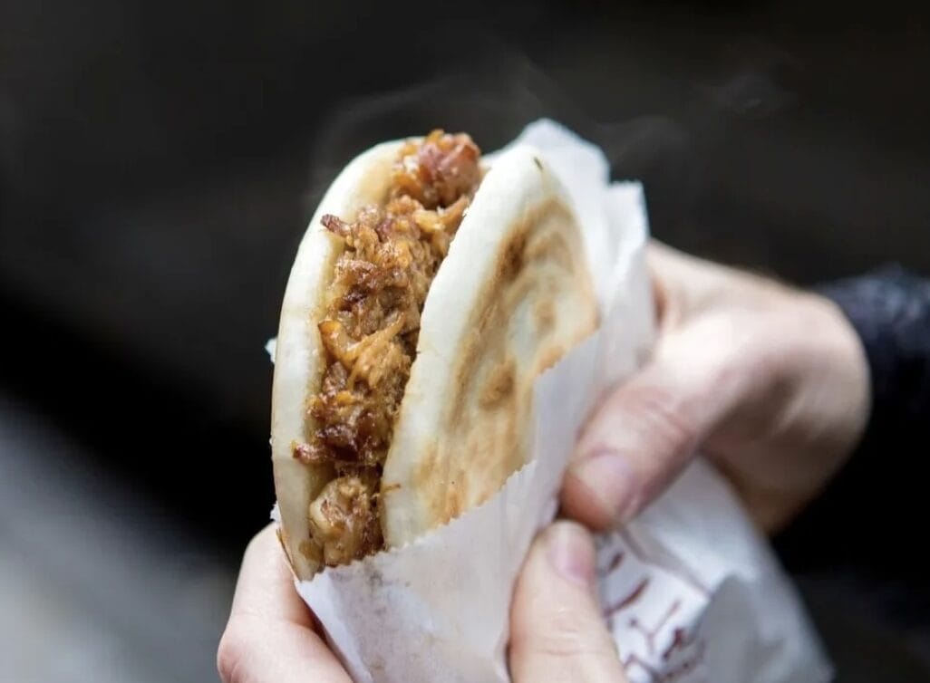 Hands hold a warm pita pocket filled with shredded meat, wrapped in paper. Steam rises from the sandwich, suggesting it’s hot.