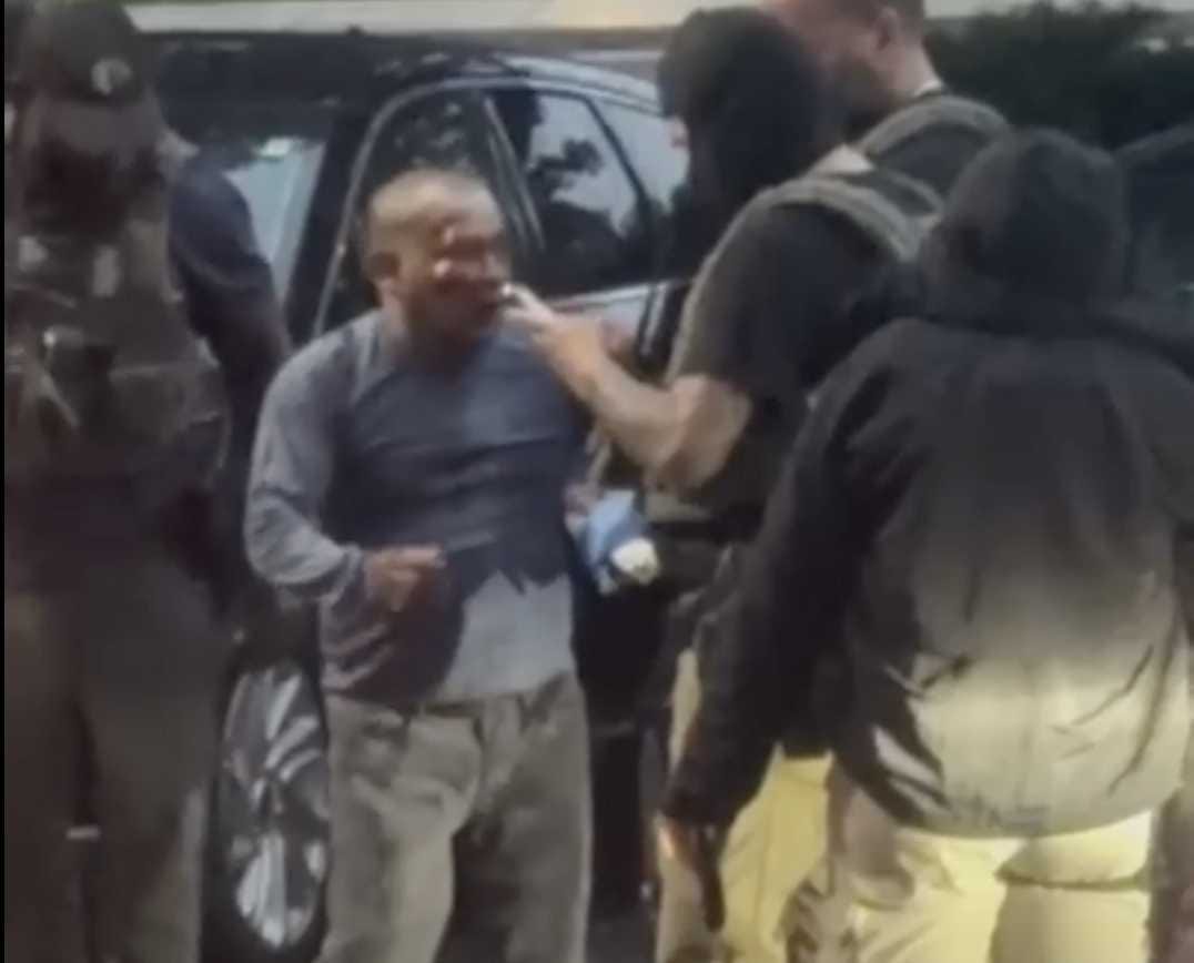 Man standing by an open car door as several armed officers in tactical gear surround him.