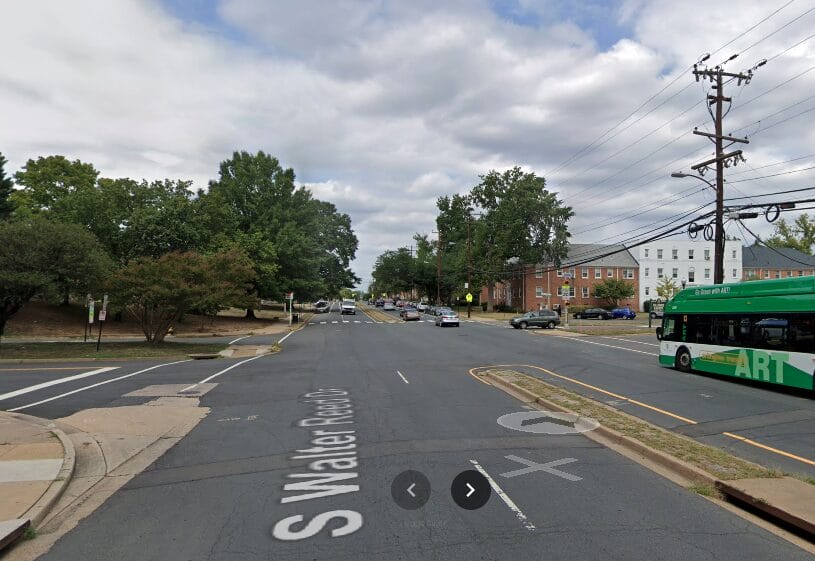 Daytime street view of a suburban intersection with a green-and-white bus on the right, brick buildings, power lines, and trees in the background.